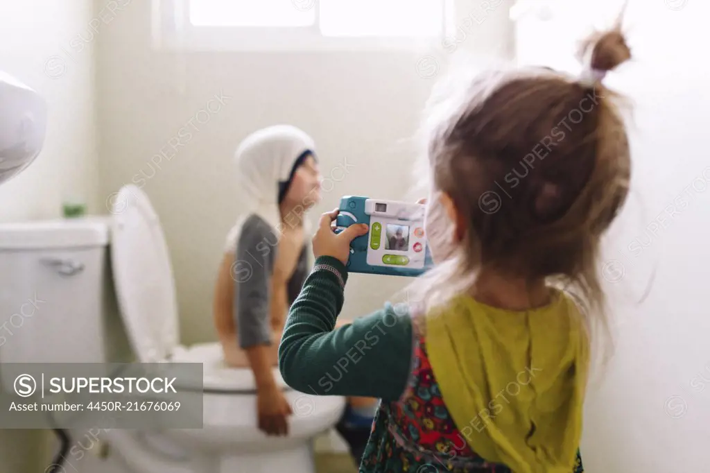 Young girl standing in bathroom, holding camera, taking picture of boy sitting on toilet with his shirt over his head.
