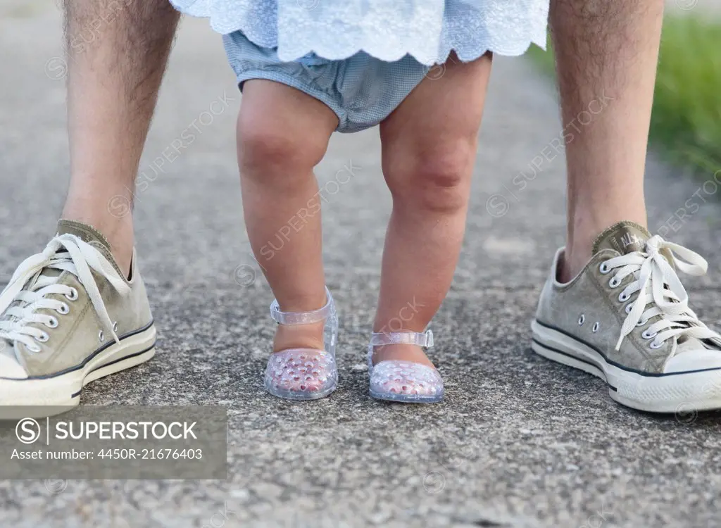 Feet of a baby girl standing between the feet of an adult, parent and child.