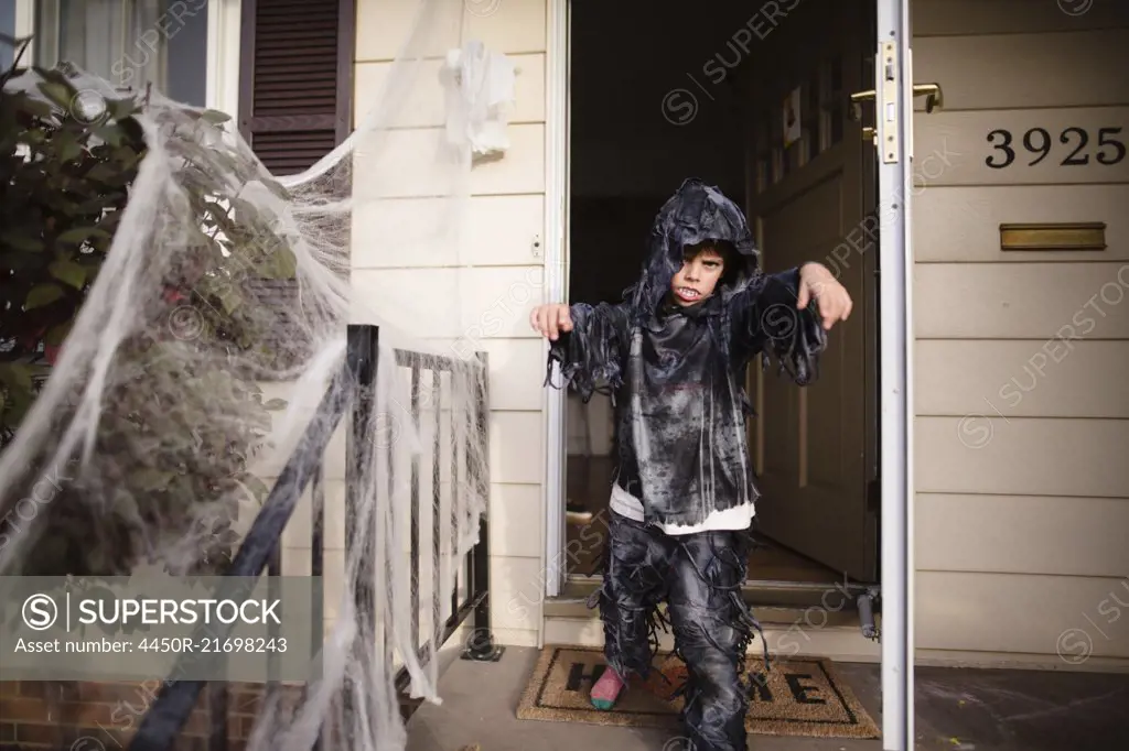 Boy wearing hooded Halloween costume standing outside front door of house, banister decorated with white netting.