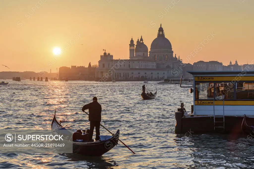 Two gondoliers on the Canale Grande in Venice, Italy, at sunrise, with the dome of Santa Maria della Salute in the distance.