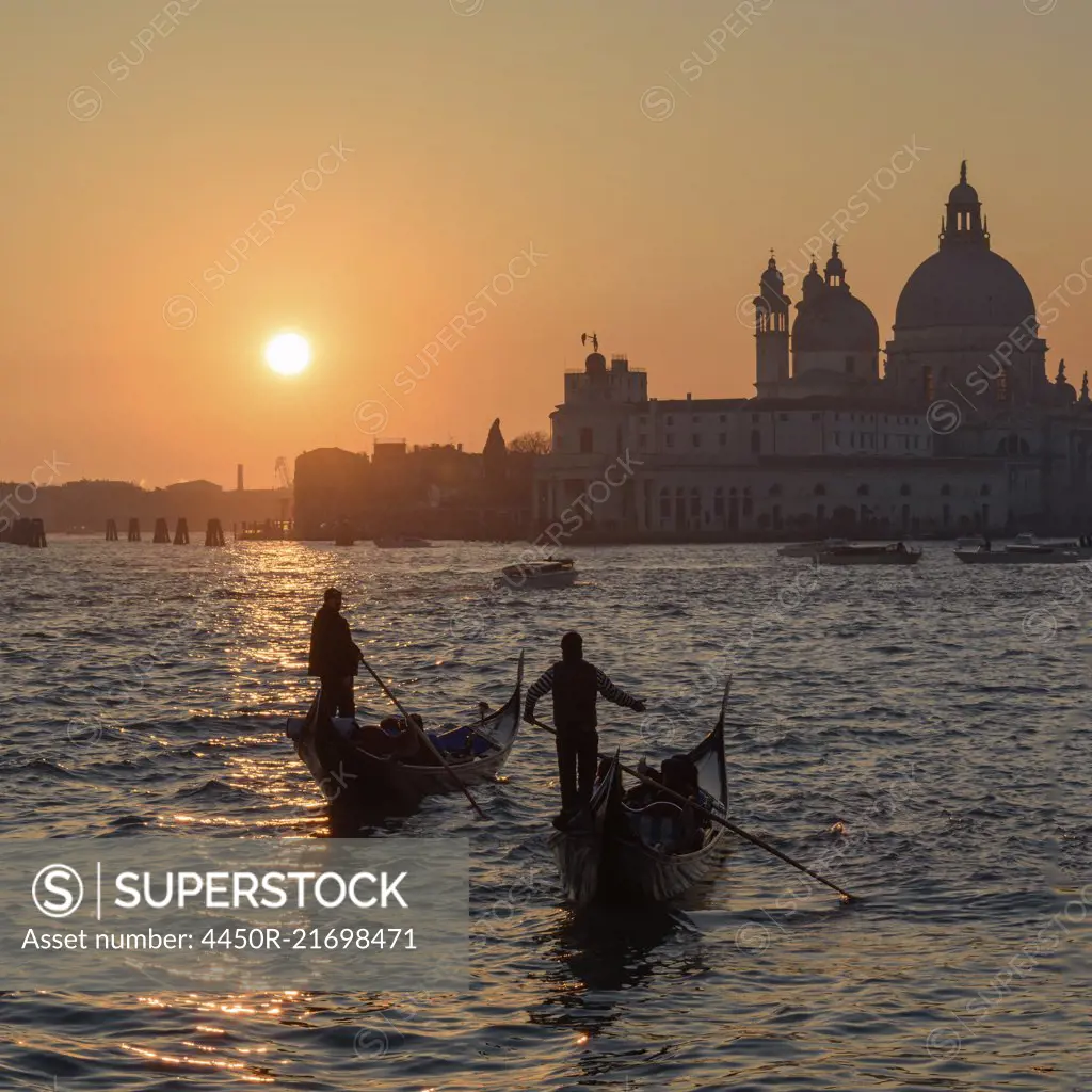 Two gondoliers on the Canale Grande in Venice, Italy, at sunrise, with the dome of Santa Maria della Salute in the distance.