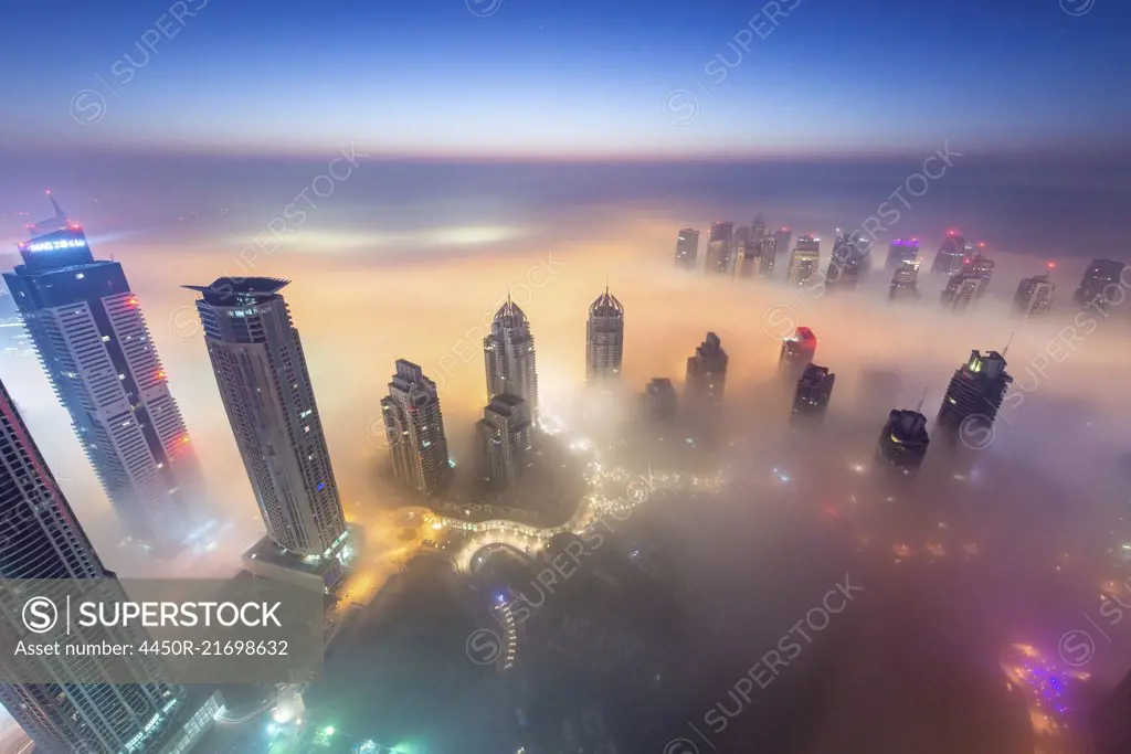 Aerial view of cityscape with illuminated skyscrapers above the clouds in Dubai, United Arab Emirates at dusk.