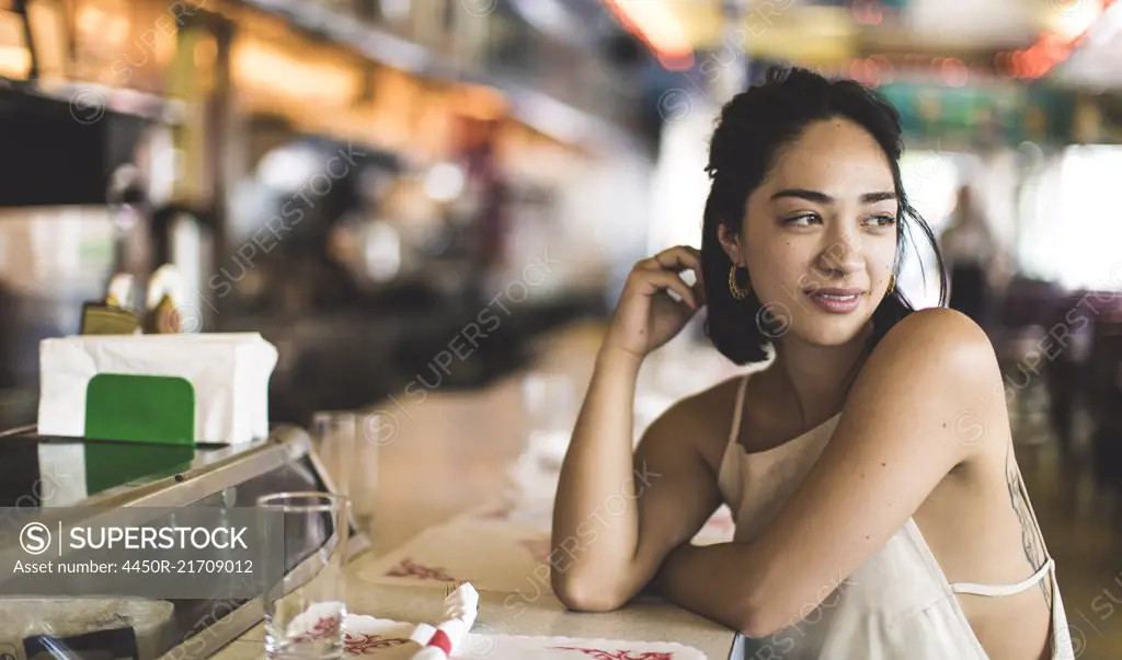 Young woman sitting at a bar counter looking over her shoulder, hand in her hair.