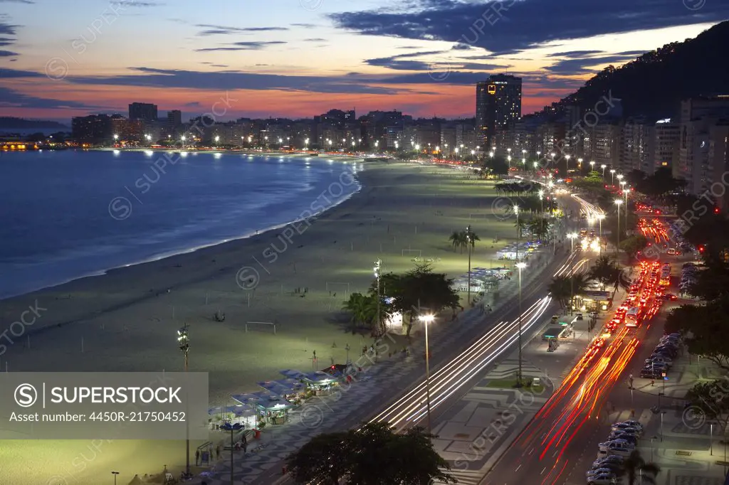 High angle view of long sandy beach lined by illuminated tall buildings, light trail of cars on avenue.