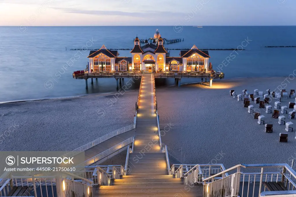 Wooden stairway and walkway leading across sandy beach towards illuminated building on a pier.