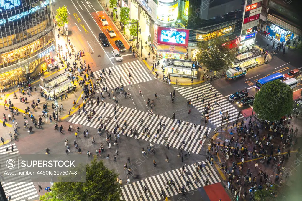 High angle view of urban street lined with illuminated buildings, pedestrians walking on pedestrian crossings.