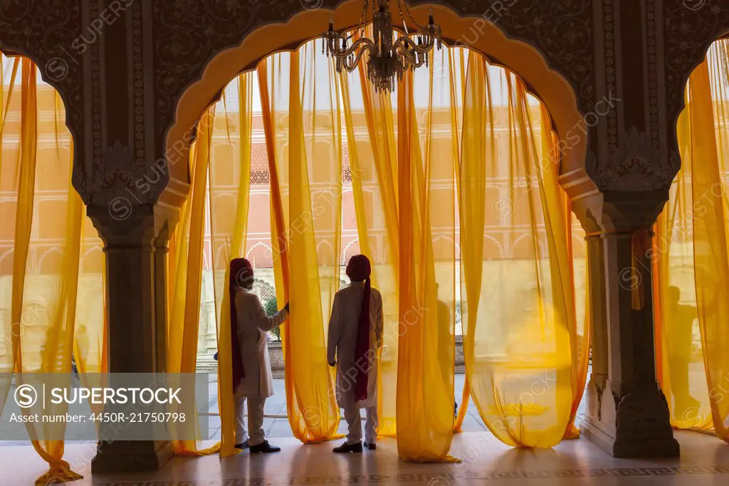 Two men wearing traditional clothing and turbans standing under arch decorated with translucent orange curtains.