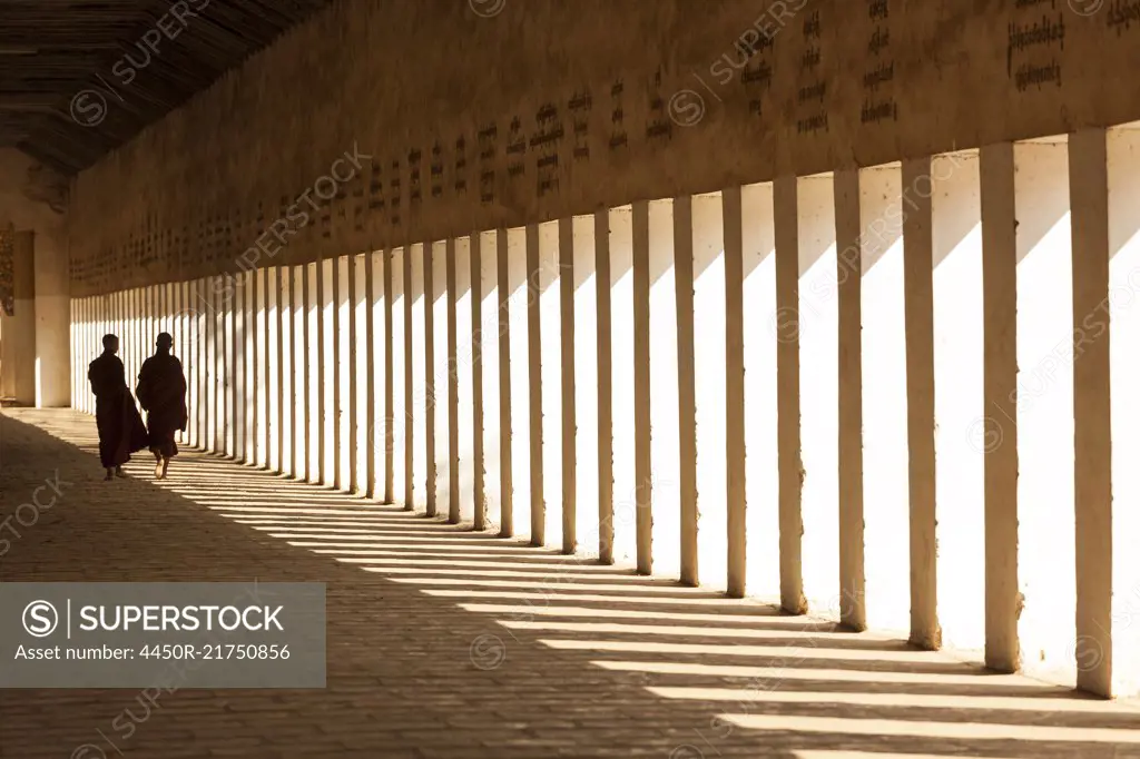 Rear view of two monks walking along sunlit colonnade of pagoda.