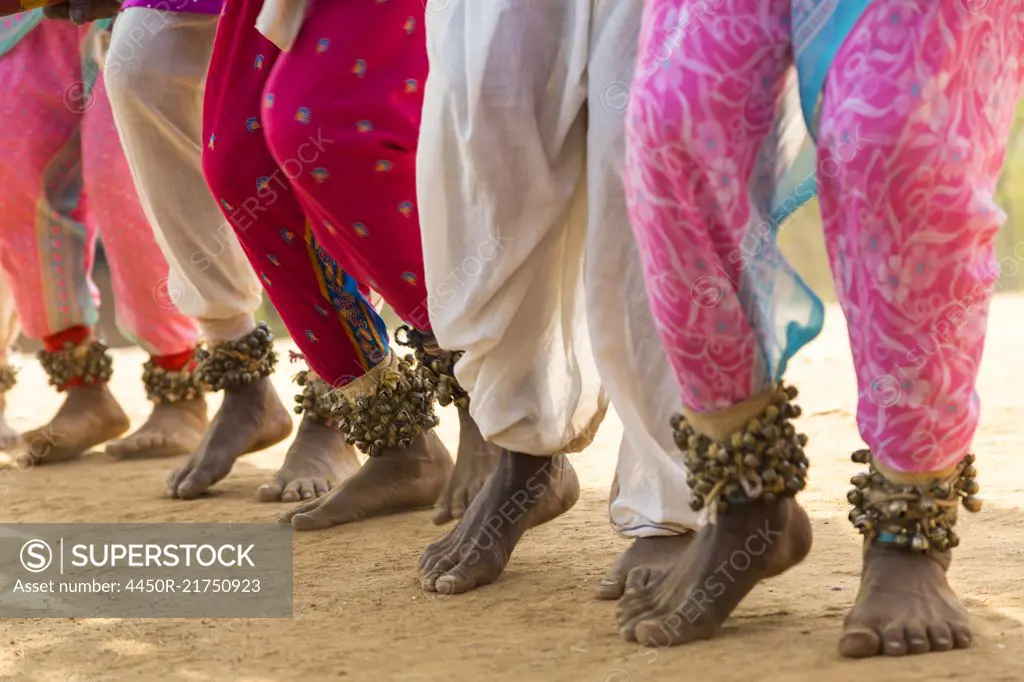 Low section view of barefoot dancers in a row on a street, wearing pink trousers and ankle bells.