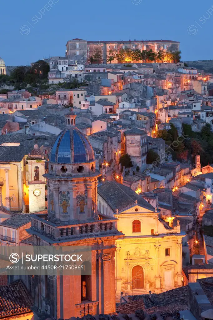 High angle view over illuminated rooftops of traditional houses in a Mediterranean city at dusk, church cupola in the foreground.