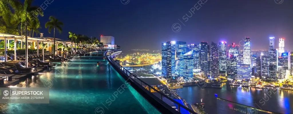 Evening view along illuminated infinity pool on roof terrace, cityscape with skyscrapers in the distance.