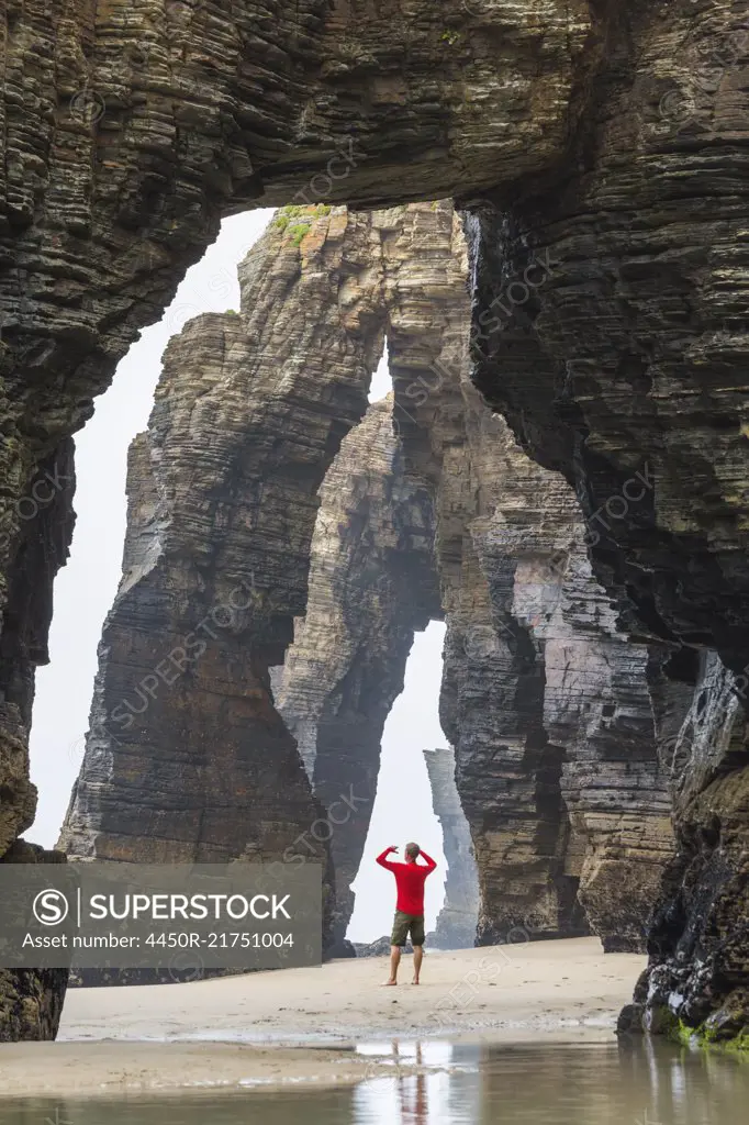 Rear view of man wearing red top standing underneath natural rock arch on sandy beach.