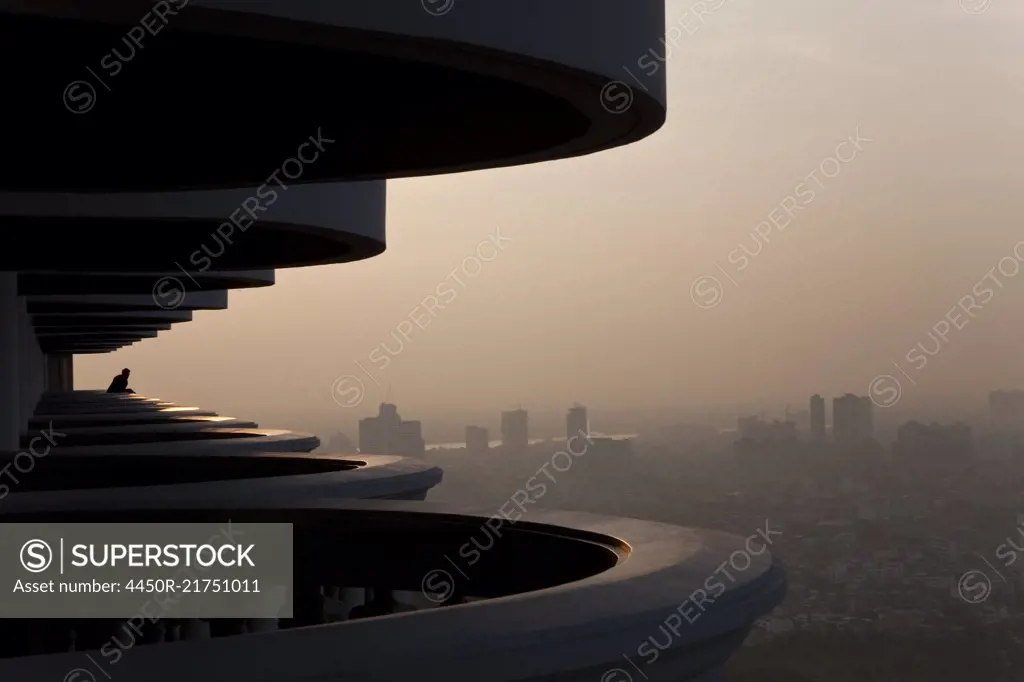 Man standing on curved balcony on skyscraper, looking over misty cityscape.