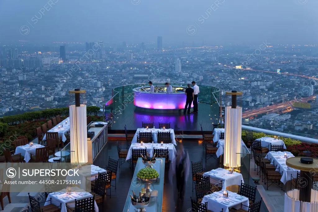 High angle view of rooftop restaurant on a skyscraper, illuminated cityscape in the distance.