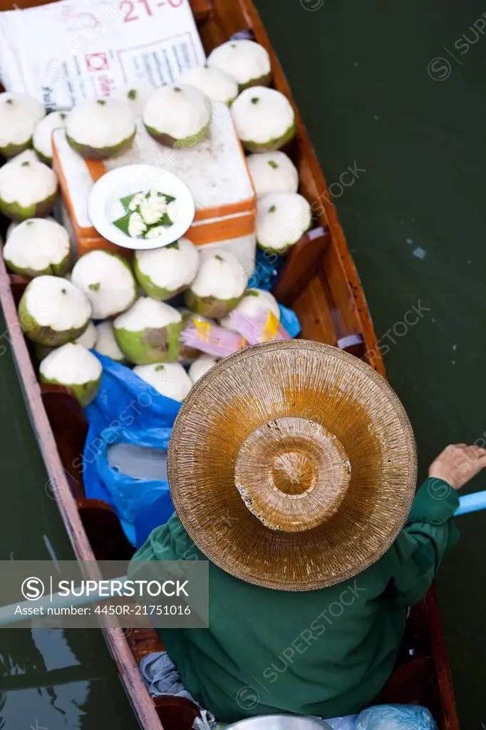 High angle view of person wearing hat sitting in narrow boat, transporting green coconuts.