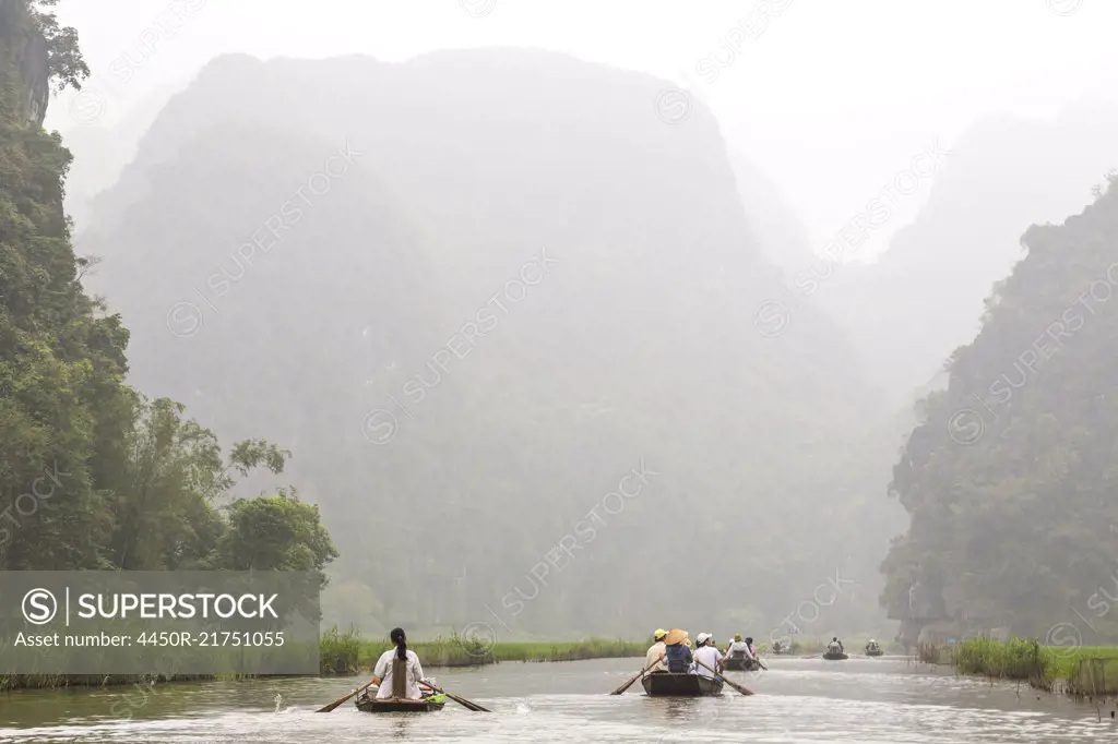 Groups of people in rowboats on a river, mountains in the background.