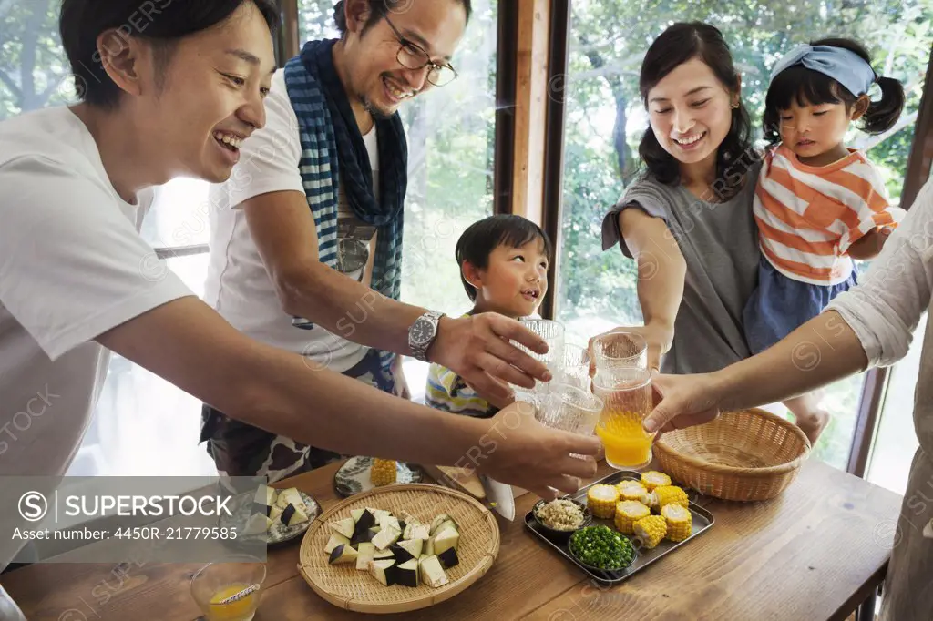 Two men, woman holding young girl and boy gathered around a table with food, holding drinking glasses, toasting.