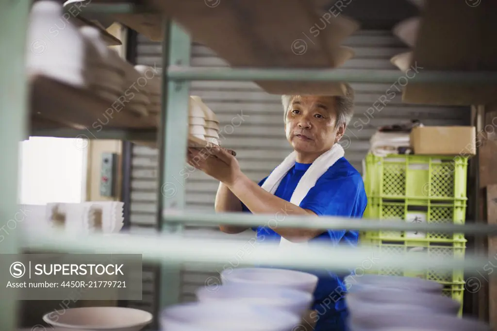 Man standing in a Japanese porcelain workshop, placing large wooden trays with white bowls onto shelf.