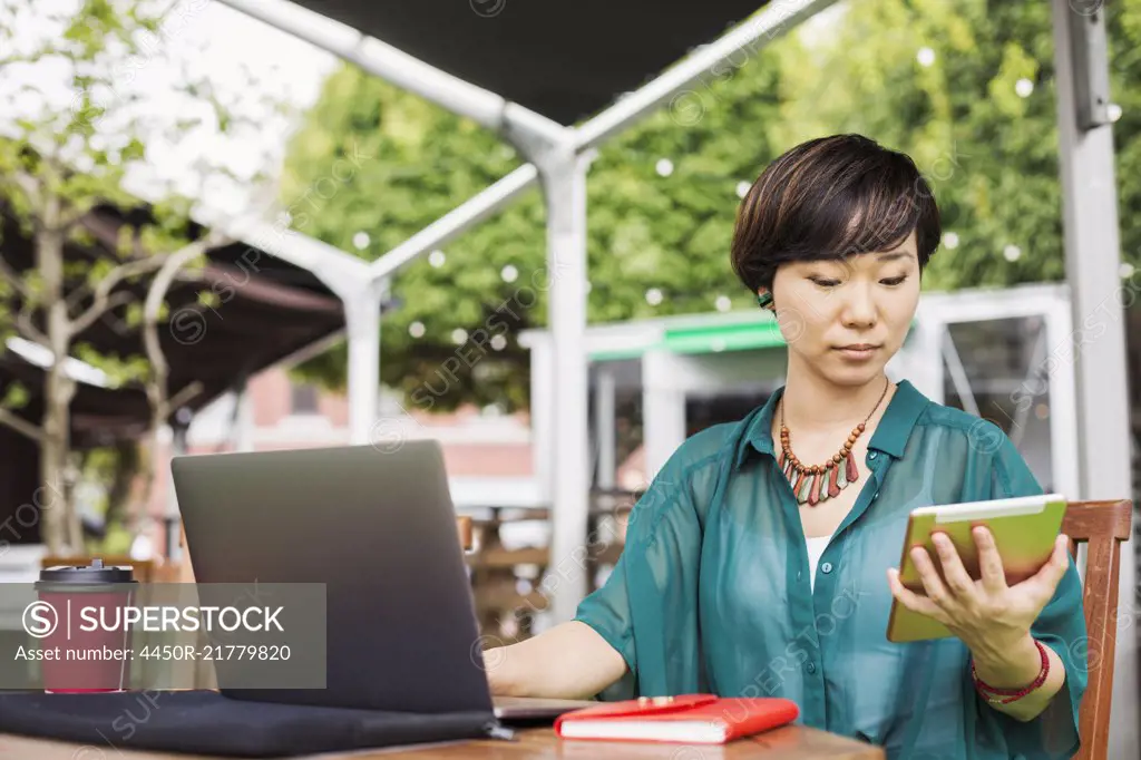 Woman with black hair wearing green shirt sitting in front of laptop at table in a street cafe, holding digital tablet.