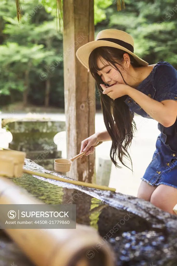 Young woman wearing blue dress and hat using bamboo water hand washing basins at Shinto Sakurai Shrine, Fukuoka, Japan.