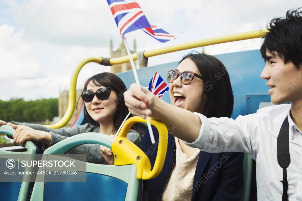 Smiling man waving Union Jack flag and two women with black hair sitting on the top of an open Double-Decker bus.