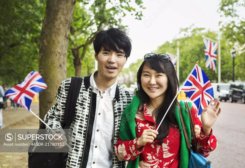 Smiling man and woman with black hair standing on the side of a tree-lined urban road, holding small Union Jack flags, looking at camera.