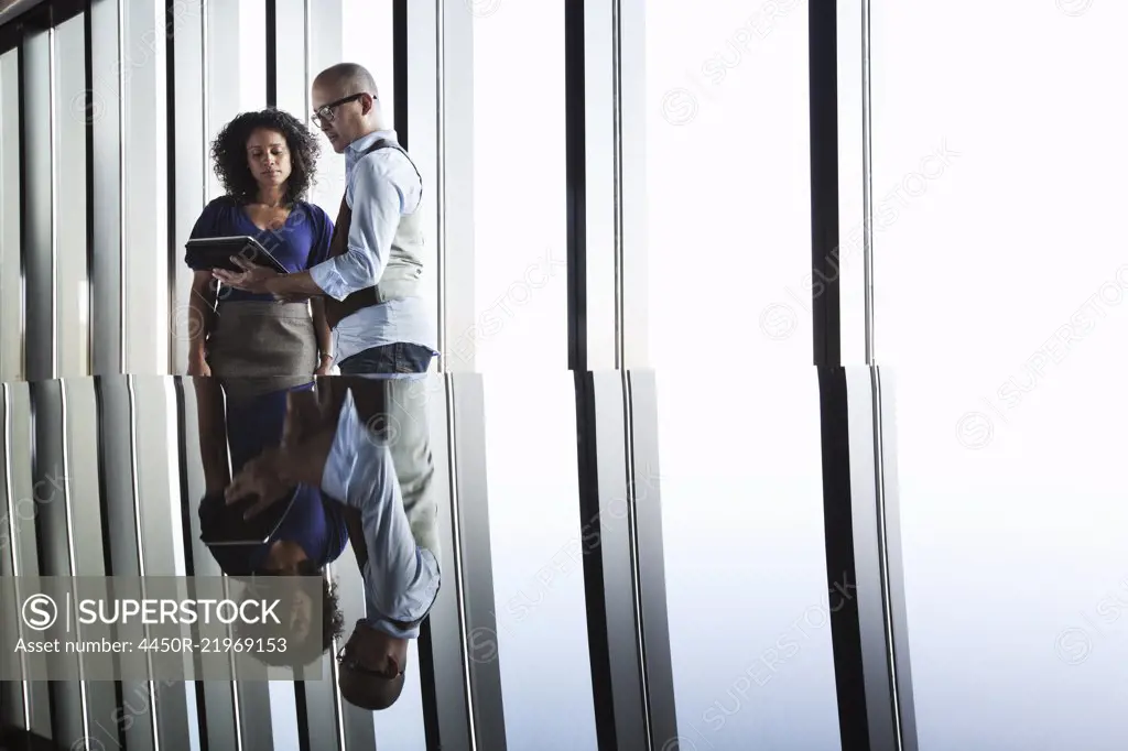 Black businesswoman and Asian businessman looking at a notebook computer and reflected on the glass top of an office desk in front of a bank of windows.