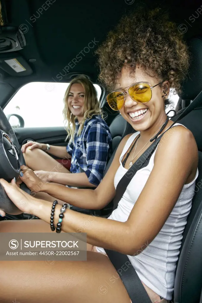 Portrait of two women with long blond and brown curly hair sitting in car, wearing sunglasses, smiling.