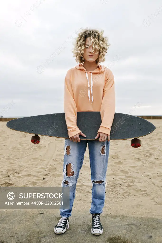 Young woman with curly blond hair wearing pink hoodie and ripped jeans standing on sandy beach, holding skateboard, looking at camera.