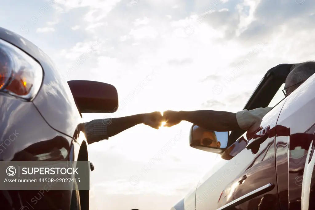 Fist bump between a passenger in one car and driver in another while cars are parked at a rest stop.