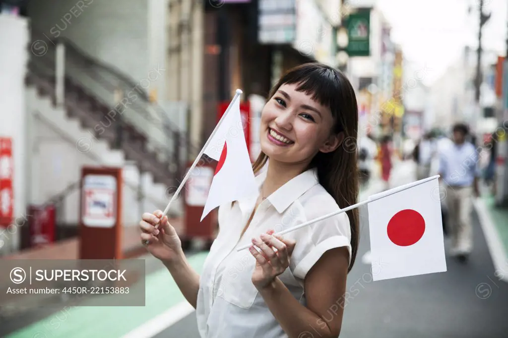 Smiling Japanese woman with long brown hair wearing white short-sleeved blouse standing in a street, holding small Japanese flag.