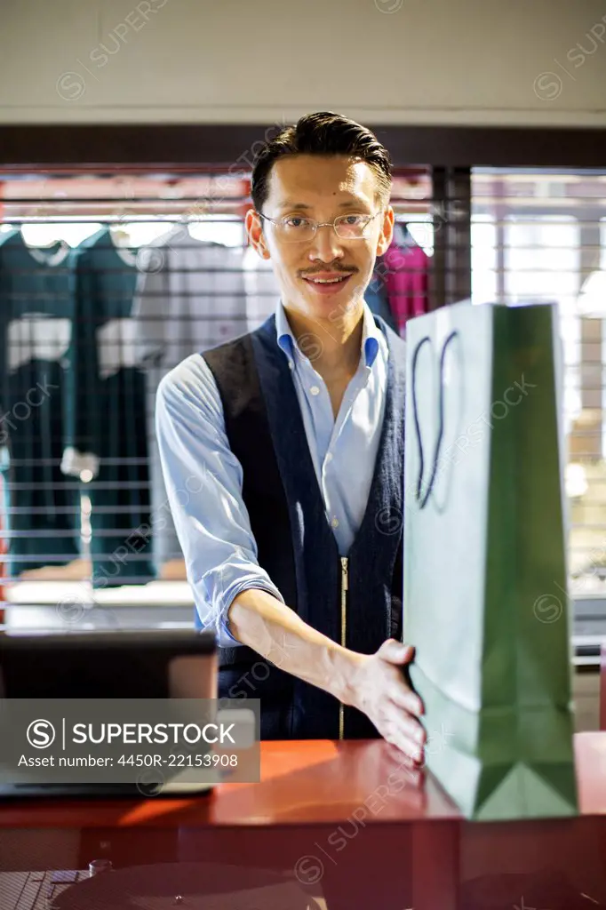 Japanese salesman with moustache wearing glasses standing at counter in clothing store, holding green shopping bag, smiling at camera.