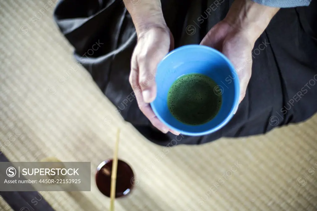 High angle close up of Japanese man wearing traditional kimono kneeling on floor holding blue bowl with Matcha tea during tea ceremony.