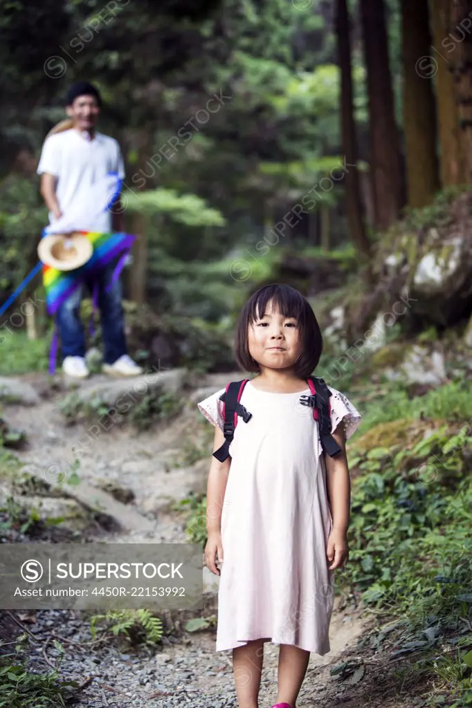 Japanese girl wearing pale pink sun dress and carrying backpack standing in a forest, man in the background.