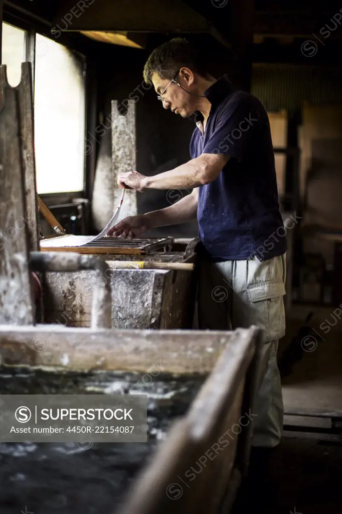 Japanese man in a workshop, holding a wooden frame with pressed pulp, making traditional Washi paper.