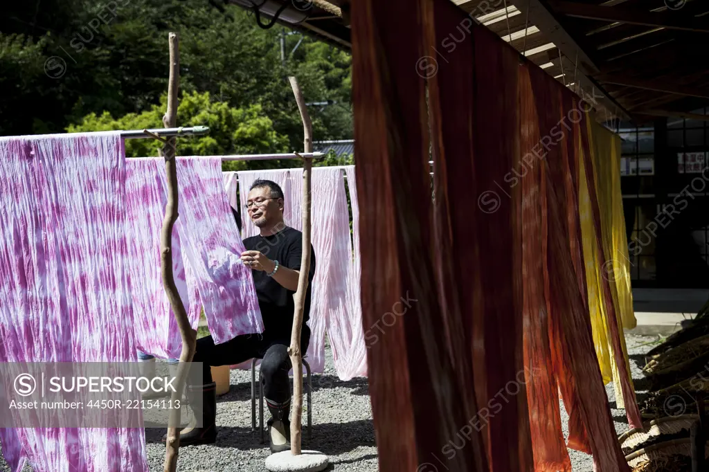 Japanese man sitting outside a textile plant dye workshop, hanging up freshly dyed pink fabric.