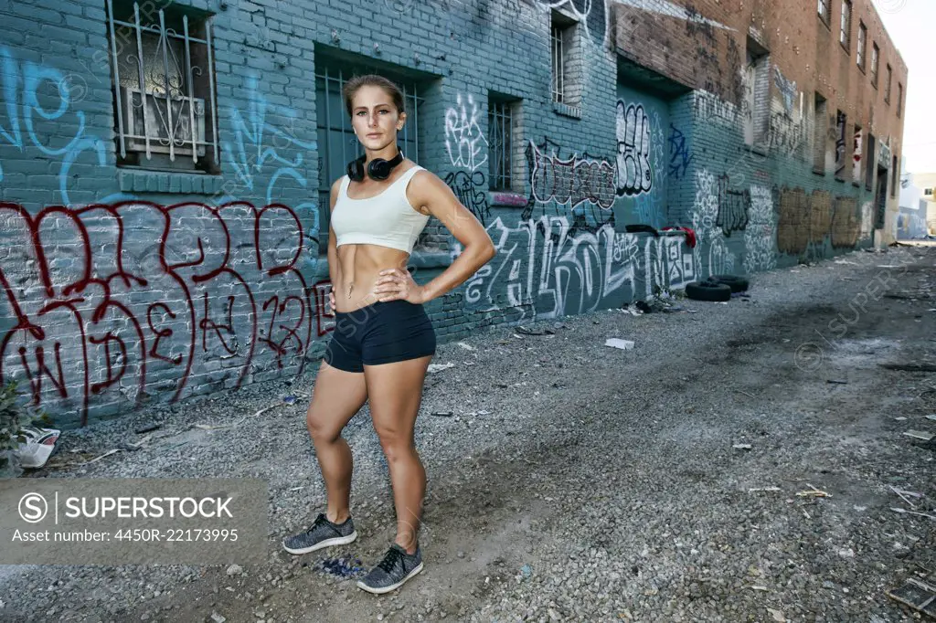 Female athlete standing on street in front of blue building covered in graffiti, looking at camera.