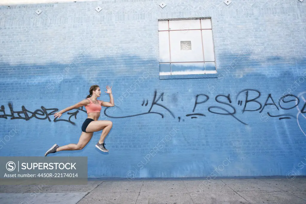 Female athlete running along sidewalk past blue building covered in graffiti.