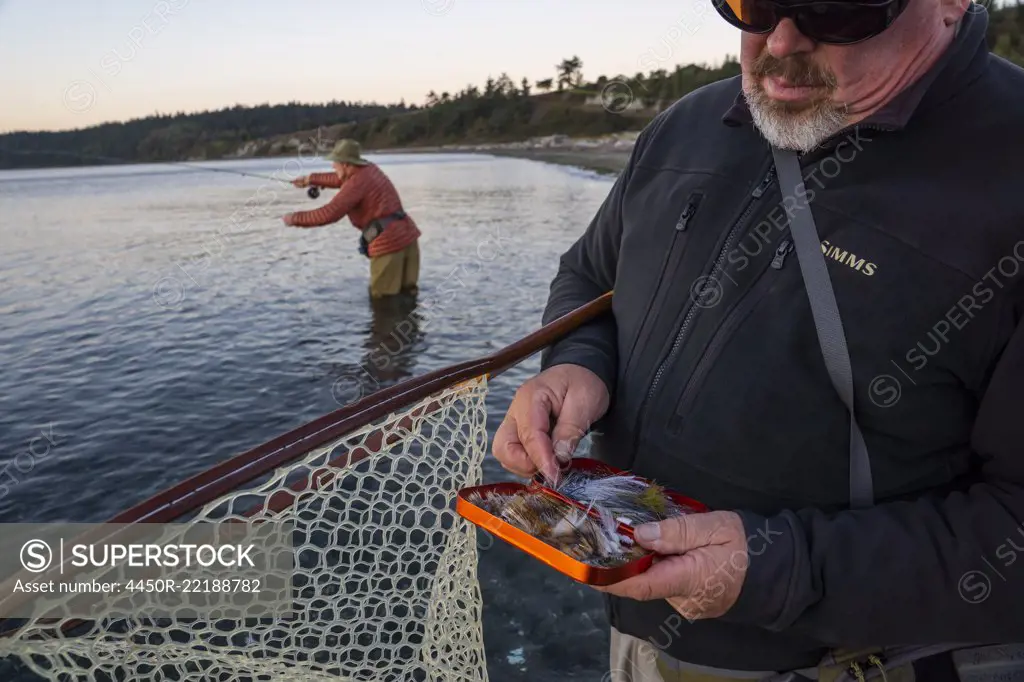 A fly-fishing guide checks his fly assortment for salmon and searun coastal cutthroat trout while his client fly fishes in the background.