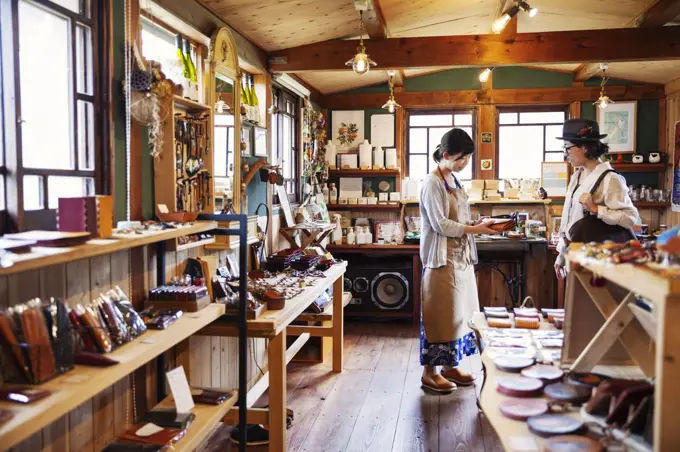 Japanese woman wearing apron standing in a leather shop, holding tan leather clutch bag, talking to customer.