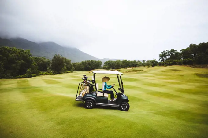 Woman wearing traditional straw hat driving golf cart on the green of a golf course under a cloudy sky.