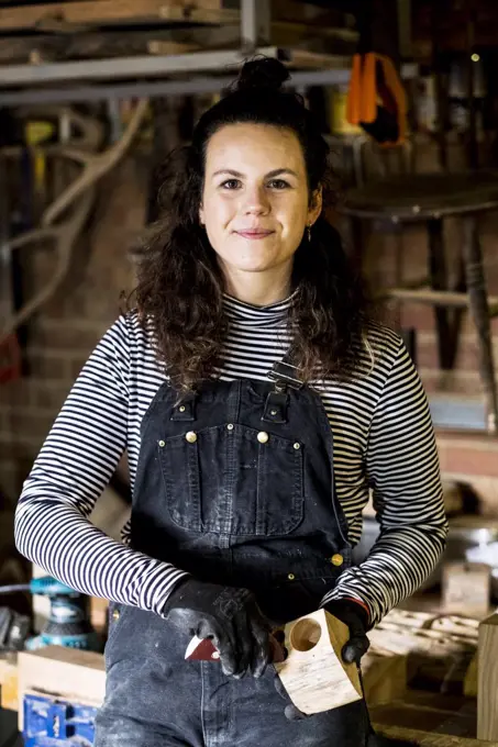 Woman with long brown hair wearing dungarees standing in wood workshop, smiling at camera.