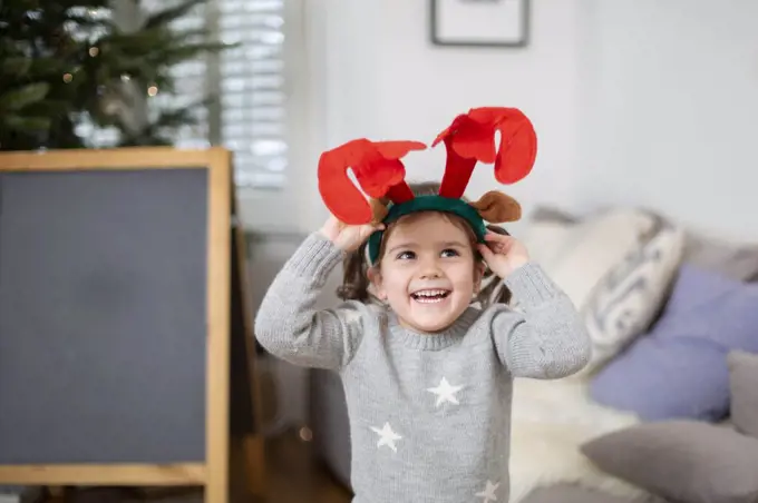 Smiling young girl wearing grey jumper putting on reindeer antler headband.