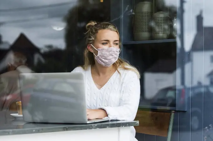 Woman wearing face mask sitting alone at a cafe table with a laptop computer, working remotely.