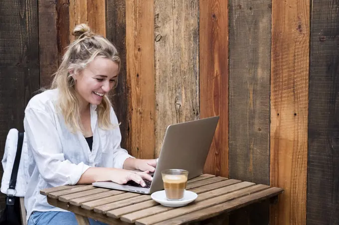 Young blond woman sitting alone at a cafe table with a laptop computer, working remotely.