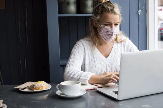 Woman wearing face mask sitting alone at a cafe table with a laptop, working remotely.