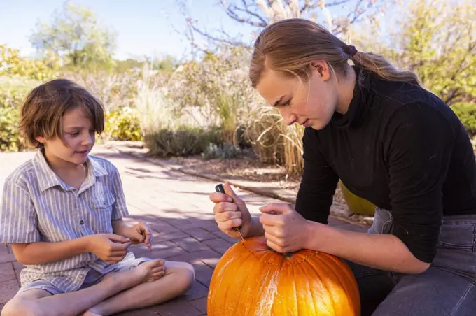 Teenage girl and her younger brother carving pumpkins on patio.