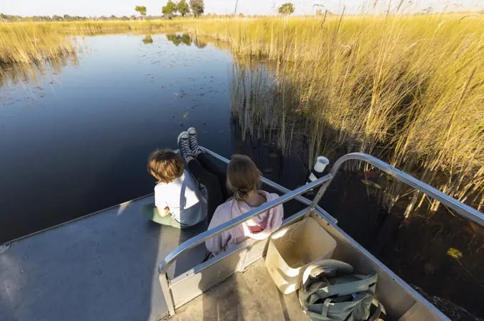 Two children seated on the stern of a boat flowing along a quiet waterway. 