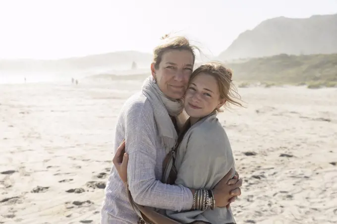 Adult woman and her teenage daughter hugging, standing on a windswept beach