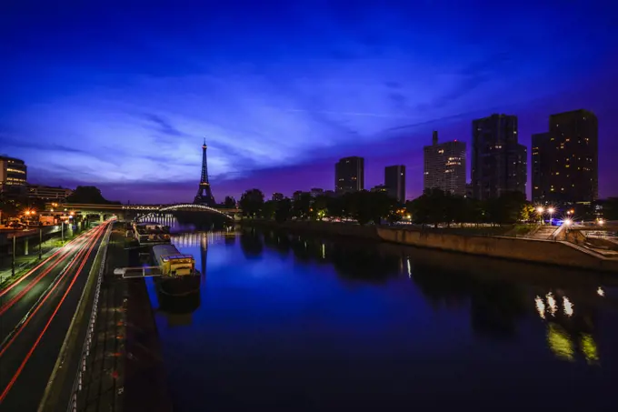 A view along the water of the River Seine at night, tall buildings on the river bank, the Eiffel tower in the distance. 